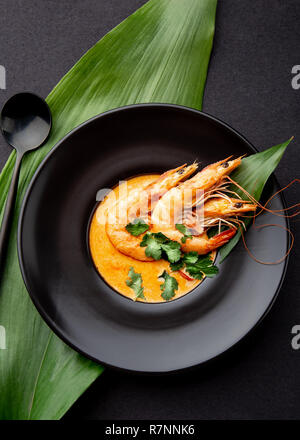 Meeresfrüchtesuppe mit ganzen Garnelen und tropische Blätter auf schwarze Platte eingerichtet, schwarzer Hintergrund Stockfoto