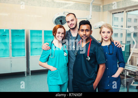 Gruppe junge Multirassischen Medizinstudenten mit Chirurgen umarmen, mit einem Lächeln in die Kamera. Krankenhaus OP-Saal in der backgroun Stockfoto