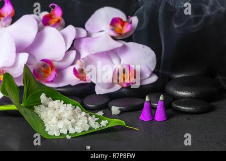 Spa-Konzept von lila Orchideen (Phalaenopsis) mit Tropfen, Aroma Räucherkegel und Meersalz auf Blatt über schwarzen zen Steine Stockfoto