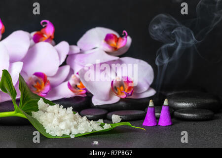 Spa Noch leben von Meersalz auf Blatt, lila Orchideen (Phalaenopsis) mit Tropfen und brennenden Aroma Räucherkegel über schwarzen zen Steine Stockfoto