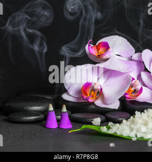 Wunderschöne Spa Konzept der lila Orchidee mit Tropfen, Aroma Räucherkegel mit Rauch- und Meersalz auf Blatt über schwarzen zen Steine Stockfoto