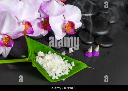 Spa Zusammensetzung der lila Orchidee mit Tropfen, Aroma Räucherkegel und Meersalz auf Blatt über schwarzen zen Steine Stockfoto
