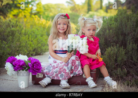 Schwestern zwei beide blond mit tiefem Eis blauen Augen tragen bunte Kleider im Central Park Wald Wiese sitzen auf Reisen trunk Bag mit Bou posing Stockfoto