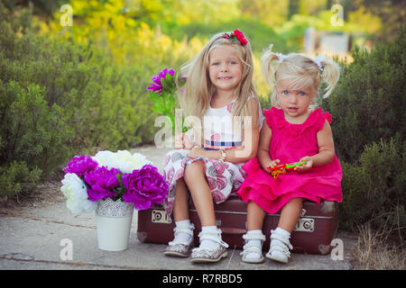 Schwestern zwei beide blond mit tiefem Eis blauen Augen tragen bunte Kleider im Central Park Wald Wiese sitzen auf Reisen trunk Bag mit Bou posing Stockfoto