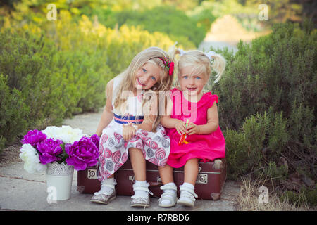 Schwestern zwei beide blond mit tiefem Eis blauen Augen tragen bunte Kleider im Central Park Wald Wiese sitzen auf Reisen trunk Bag mit Bou posing Stockfoto