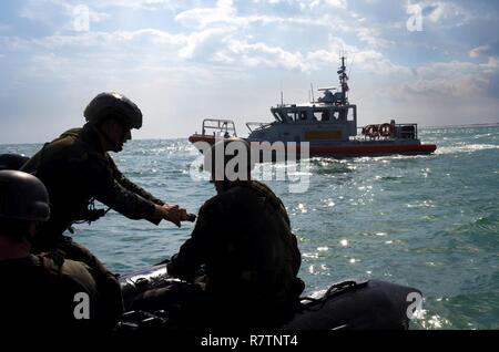 Ein Marine mit Hotel Company, 2 Raider Bataillon, Marine Corps Forces Special Operations Command (MARSOC) in Camp Lejeune in North Carolina, startet der Motor eines Zodiac starre Hülle Schlauchboot, während ein 45-Fuß-Antwort Boat-Medium Crew von der Coast Guard Station Wrightsville Beach, North Carolina, in der Nähe wartet aus Onslow Strand auf Base, 28. März 2017. Die Küstenwache boot Mannschaftsräumen waterside unterstützen, während die Marines mehrfach vom Hubschrauber gesprungen, schwamm ans Ufer und pilotiert der Tierkreis Boote. Stockfoto