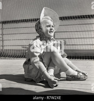 1950, historische, ein junges Mädchen mit überkreuzten Beinen auf dem Deck des Dampfers trägt ein top, rock und ein hübsches Motorhaube, ein Tuch hat unter ihr Kinn mit einem Rand über dem Gesicht gebunden, ein Zubehör allgemein in dieser Zeit getragen von Frauen und Kinder, auch wenn heute gesehen, als altmodisch sein würde. Stockfoto