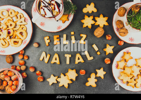 FELIZ NATAL COOKIES. Wörter Frohe Weihnachten en Portugiesisch mit gebackenen Plätzchen, Weihnachtsschmuck und Muttern auf schwarzem Schiefer Hintergrund. Weihnachtskarte für Portugiesisch sprechenden Länder der Ansicht von oben. Stockfoto