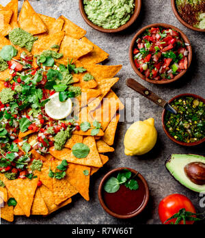 Nachos. Totopos mit Saucen. Mexikanisches Essen Konzept. Gelber Mais totopos Chips mit verschiedenen Saucen salsas-Pico del Gallo, Guacamole, Salsa verde, Paprika pebre und frischen Avocado, Tomate, Zitrone und Koriander auf Stein grauer Hintergrund, Ansicht von oben Stockfoto