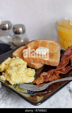 Schwarze Platte mit Rührei, Toast und Speck. Portrait Nahaufnahme. Platte positioniert wie ein Diamant. Salz und Pfeffer abschmecken und Orangensaft in den hinterg Stockfoto