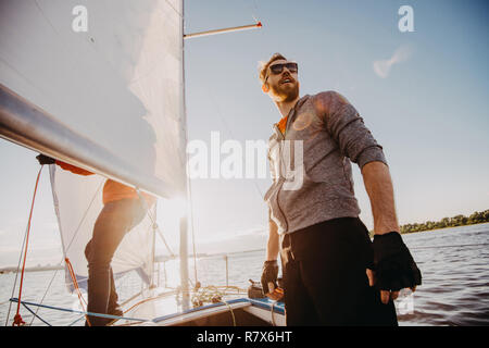Low Angle Shot eines jungen Mannes Seemann mit Sonnenbrille und Handschuhe auf einem Segelboot bei Sonnenuntergang. Bild Farben in warmen Farben. Stockfoto