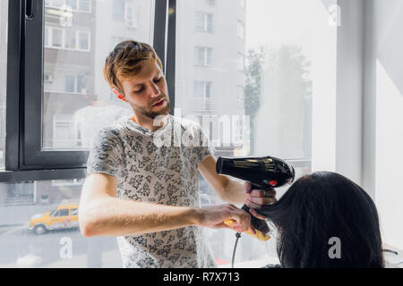 Professionelle smiling männlichen Stylist föhnen Woman's Haar mit einem Trockner im Salon Stockfoto