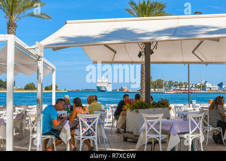 Touristen genießen Mittagessen in einem Café Waterfront in der Hafenstadt Brindisi Italien als massive Kreuzfahrt Schiff in einen Hafen geschleppt. Stockfoto