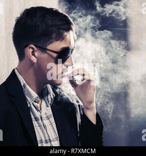 Jungen gutaussehenden Mann in Sonnenbrille Rauchen einer Zigarette in der city street Stilvolle Mode männliches Modell in Schwarz Blazer Stockfoto
