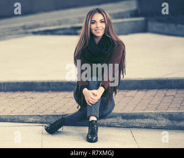 Glückliche junge Frau sitzt auf dem Bürgersteig in der Straße der Stadt. Stilvolle Mode Modell in Leder fransen Wildleder Jacke und blaue Jeans Stockfoto