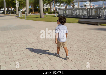 Eine selbstbewusste kleine Junge mit Sonnenbrille geht weg von den Docks an einem sonnigen Sommertag. Im Tennis Schuhe, khaki Hosen gekleidet, und blau gestreiften Hemd. Stockfoto