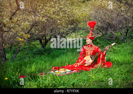 Kasachischen Frau in Rot Kostüm spielen dombra auf dem grünen Rasen in der Apple Garten von Almaty, Kasachstan, Mittelasien Stockfoto