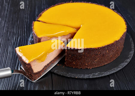 In Scheiben geschnittene Schokolade Mango cheesecake. ein Stück auf einem Kuchen Schaufel auf einen schwarzen Stein Platte auf einem schwarzen Holz- Tabelle, Ansicht von oben, close-up Stockfoto