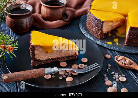 Ein Stück Schokolade Mango cheesecake serviert auf einem schwarze Platte mit Dessert Gabel und Chocolate Chips. Tanne und Tassen mit Kaffee auf einem Holztisch Stockfoto