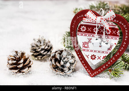 Verzierte hölzerne Herz mit weihnachtlichen Motiven gegen eine Tanne Zweig neben einem tannenzapfen auf dem Schnee lehnend. Stockfoto