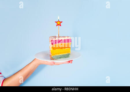 Weibliche Hand, die eine weiße Platte mit Slice von Rainbow Kuchen mit birning Kerze in der Form von Sternen auf blauem Hintergrund isoliert. Happy bithday, party Stockfoto