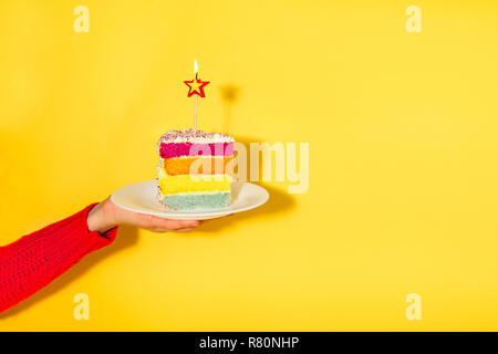 Weibliche Hand, die eine weiße Platte mit Slice von Rainbow Kuchen mit birning Kerze in der Form von Stern auf gelbem Hintergrund isoliert. Happy bithday, Teil Stockfoto
