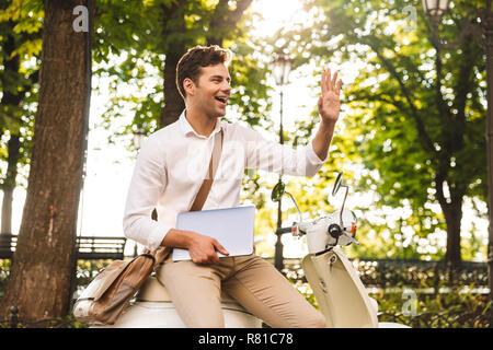 Fröhlicher junger Geschäftsmann sitzen auf einem Motorrad draußen, Laptop Stockfoto