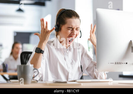 Wütende junge Frau mit Mikrofon Headset im Hemd an Ihrem Arbeitsplatz im Büro sitzen, gekleidet, schreien Stockfoto