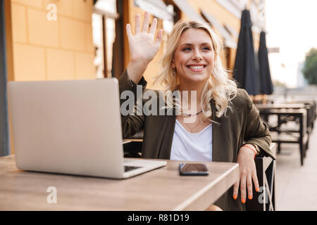 Portrait von lächelnden blonde Frau tragen Jacke mit silber Notebook und Smartphone beim Sitzen im Cafe im Freien Stockfoto