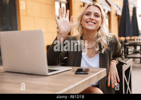 Portrait von Fröhliche blonde Frau tragen Jacke mit silber Laptop und Smartphone, während im Cafe sitzen im Freien Stockfoto