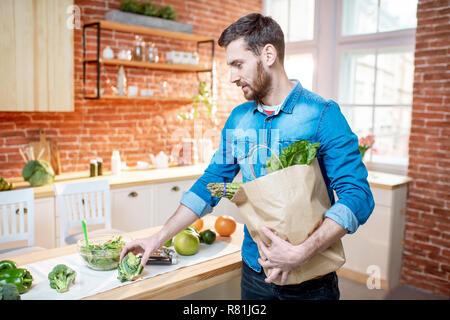 Schöner Mann im blauen Hemd stehend mit Einkaufstasche voller gesunder Grün essen in der Küche Stockfoto