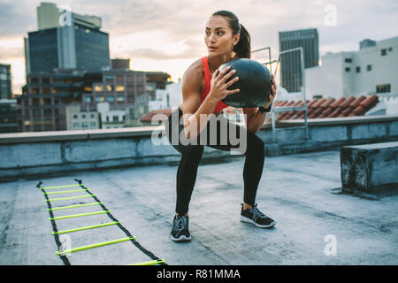 Fitnesstraining-Frau auf dem Dach mit einem Medizinball. Frau, die Squats macht, hält einen Medizinball mit einer Agilitätsleiter an ihrer Seite auf dem Dach Stockfoto