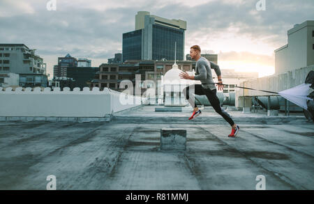 Seitenansicht eines Fitness Mann laufen auf der Dachterrasse mit Widerstand Fallschirm. Athlet Training auf der Dachterrasse mit einem Fallschirm zu seiner Taille gebunden. Stockfoto