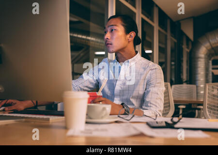 Asiatische Geschäftsmann an seinem Schreibtisch arbeiten am Computer sitzen spät in der Nacht mit Kaffee und Essen auf dem Tisch. Man ernsthaft arbeiten im Büro auf Stockfoto