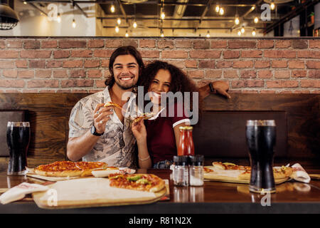 Portrait von glückliches junges Paar zusammen sitzen im Restaurant mit Pizza in der Hand. Lächelnd Mann und Frau im Cafe sitzen mit Pizza auf dem Tisch. Stockfoto