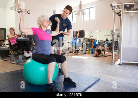 Ältere Frau Ausübung auf Schweizer Kugel mit Gewichten wird gefördert vom persönlichen Trainer im Fitnessraum Stockfoto