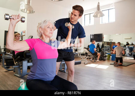 Ältere Frau Ausübung auf Schweizer Kugel mit Gewichten wird gefördert vom persönlichen Trainer im Fitnessraum Stockfoto