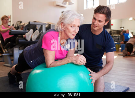 Ältere Frau Ausübung auf Schweizer Kugel durch Personal Trainer im Fitnessstudio gefördert Stockfoto