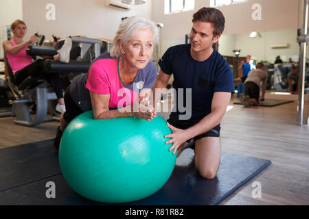 Ältere Frau Ausübung auf Schweizer Kugel durch Personal Trainer im Fitnessstudio gefördert Stockfoto