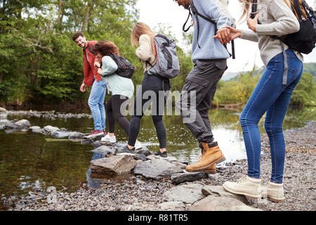 Multi-ethnische Gruppe von fünf jungen Erwachsenen Freunde halten Hände wandern auf den Felsen einen Stream bei einer Wanderung zu überqueren, Nahaufnahme, Ansicht von hinten Stockfoto