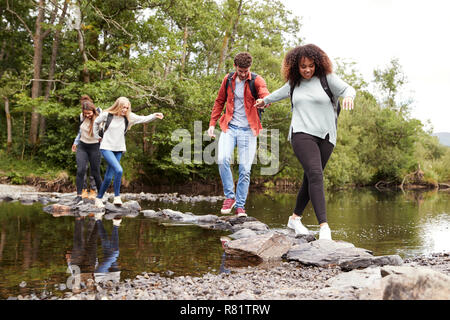 Fünf gemischten Rennen junge erwachsene Freunde halten Sie Hände und einander helfen, während sorgfältig über einen Stream auf Steine während einer Wanderung Stockfoto