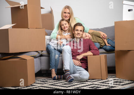 Bild der glücklichen Mann, Frau, Mädchen und junge sitzt auf grau Sofa unter Kartons für Umzug in neue Wohnung Stockfoto