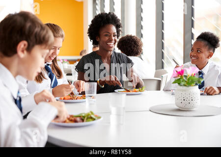 Lehrerin mit einer Gruppe von Schülerinnen und Schüler tragen Uniform Sitzen um den Tisch und Essen Mittagessen in der Cafeteria Stockfoto