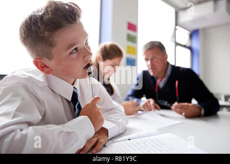 Die Konzentration männlicher High School Student trägt Uniform Arbeiten am Tisch mit dem Lehrer sprechen mit Schülern im Hintergrund Stockfoto