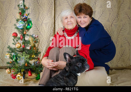 Familie Momente zur Weihnachtszeit. alte Frau lächeln. Großmutter und ihrer Tochter sitzen in der Nähe von Weihnachten Baum. Geist der Neuen Jahr. umarmen sich Stockfoto