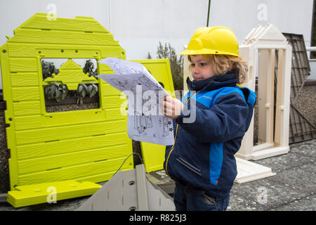 Kleiner Junge, 3 Jahre, mit Spielzeug hard hat, hilft das Spielhaus zu bauen, studieren die Blaupause Stockfoto