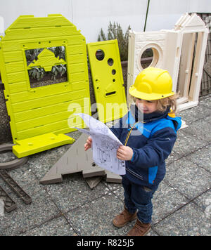 Kleiner Junge, 3 Jahre, mit Spielzeug hard hat, hilft das Spielhaus zu bauen, studieren die Blaupause Stockfoto