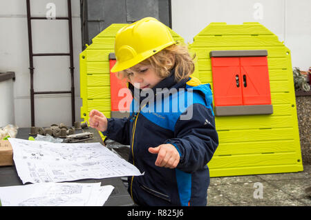 Kleiner Junge, 3 Jahre, mit Spielzeug hard hat, hilft das Spielhaus zu bauen, studieren die Blaupause Stockfoto