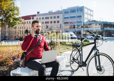 Geschäftsmann Pendler mit dem Fahrrad sitzt auf der Bank, mit Laptop und Smartphone. Stockfoto