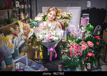Schöne und attraktive Florist ist unter bunten Blumen. Sie halten bouqette in Händen und es betrachten. Junge Frau lächelt. Stockfoto